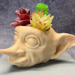 Puede incluir: Un macetero de cerámica beige con forma de cabeza de duende con orejas grandes. El macetero está lleno de suculentas artificiales en tonos verdes, rojos y amarillos.