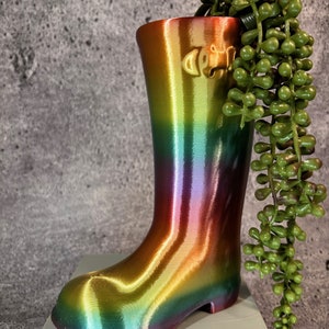 Pode incluir: Um vaso em forma de bota iridescente arco-íris com uma planta suculenta verde dentro. A bota tem um acabamento brilhante e metálico.