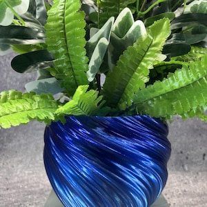 May include: A vibrant blue planter with a spiraling, ribbed texture holds a lush arrangement of artificial greenery. The plant features a mix of bright green fern-like fronds and broad, variegated leaves with white edges, creating a dense foliage display.