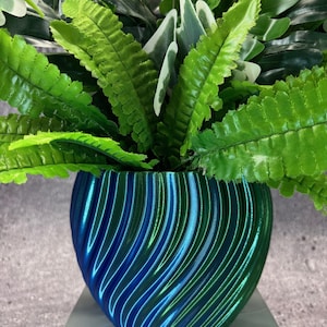 May include: A vibrant, iridescent blue and green 3D-printed planter pot with a swirling design, filled with lush green artificial ferns and other foliage. The pot sits on a light gray surface, creating a striking contrast.