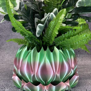 May include: A 3D printed plant pot with a scalloped, petal-like exterior featuring a gradient of metallic green and pink hues. The pot includes a matching detachable saucer base and is filled with a variety of artificial green foliage plants.