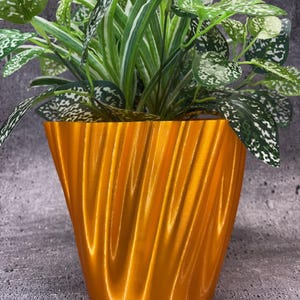 May include: A gold-colored, wavy, ceramic planter with a green leafy plant inside.