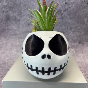 May include: A white, spherical planter featuring a black Jack Skellington face design. The planter holds a small, green and brown succulent plant. The planter is sitting on a light gray surface.