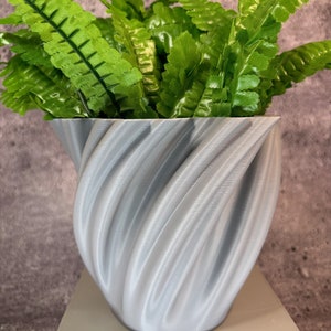 May include: A gray, 3D printed planter with a swirling design, holding a green artificial fern plant.