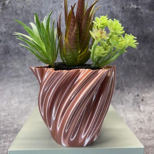 Op de afbeelding: Een decoratieve koperkleurige plantenbak met een spiraalvormig ontwerp, met drie kunstmatige vetplanten. De vetplanten zijn groen en bruin en geven de moderne, getextureerde pot een natuurlijke uitstraling. De plantenbak staat op een lichtgroen oppervlak.