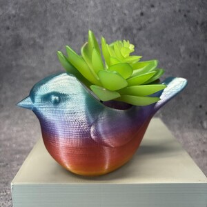Puede incluir: Una maceta con forma de pájaro con un acabado degradado iridiscente que transita del azul en la cabeza al morado y naranja en la base. Una planta suculenta artificial de color verde brillante se encuentra en la abertura superior de la figura.