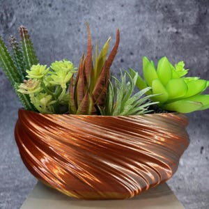 Puede incluir: Una maceta decorativa de color cobre con un diseño en espiral, llena de una variedad de suculentas artificiales. Las suculentas incluyen plantas verdes, marrones y verde claro, creando una exhibición vibrante.