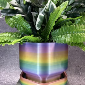 May include: A hexagonal plant pot with a matching saucer, featuring a horizontal rainbow gradient finish in shades of purple, blue, green, yellow, orange, and pink. The pot contains a lush arrangement of green and white variegated artificial foliage.
