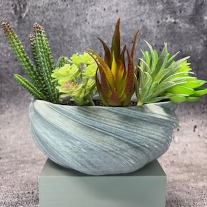 Puede incluir: Un arreglo decorativo de suculentas artificiales en un cuenco de cerámica gris con remolinos. Las suculentas incluyen cactus verdes, plantas de color amarillo verdoso y marrón rojizo. El cuenco se encuentra sobre un soporte rectangular verde claro sobre un fondo gris texturizado.