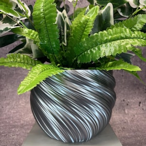 Puede incluir: Una maceta plateada con un diseño de remolino y texturado contiene una planta verde frondosa. La maceta está sentada sobre un pedestal gris.