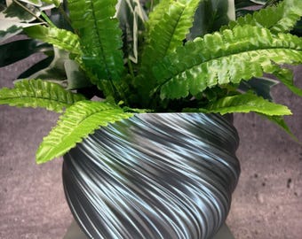 May include: A silver planter with a swirling, textured design holds a green leafy plant. The planter is sitting on a gray pedestal.