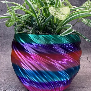 Puede incluir: Una maceta multicolor con un diseño en espiral, llena de una planta de hojas verdes y blancas. Los colores de la maceta van del verde azulado al morado, con bandas de rosa, naranja y azul. La maceta tiene un acabado texturizado y brillante.