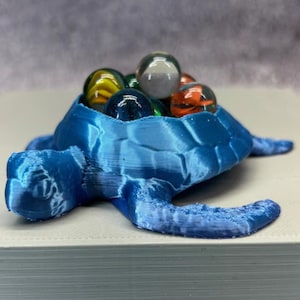 Könnte beinhalten: Ein blaues, 3D-gedrucktes, schildkrötenförmiges Gefäß, gefüllt mit bunten Murmeln. Die Schildkröte hat einen strukturierten Panzer und ausgestreckte Flossen. Die Murmeln sind in verschiedenen Farben, darunter Gelb, Orange, Klar und Dunkelblau. Der Behälter steht auf einer weißen Oberfläche.