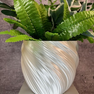 May include: A white, pearlescent, spiral-textured planter filled with vibrant green and white artificial plants. The planter sits on a small, square, light-colored pedestal, creating a decorative display. The leaves have a realistic appearance.