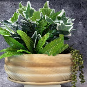 Op de afbeelding: Een decoratieve plantenbak met een lichtbruine, houtachtige afwerking, gevuld met levendige groene en witte kunstplanten. De plantenbak heeft een afgeronde, langwerpige vorm en staat op een bijpassende schotel. Groene kralen vallen van de zijkant naar beneden.