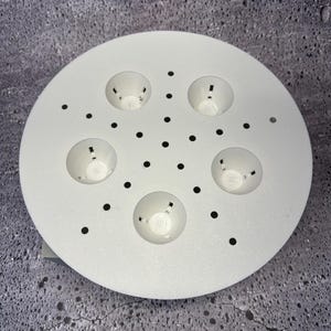 May include: A white, circular object with five recessed, bowl-shaped areas and numerous small, evenly spaced black dots. The object appears to be made of a solid, possibly metal, material. The design suggests a functional or decorative purpose.