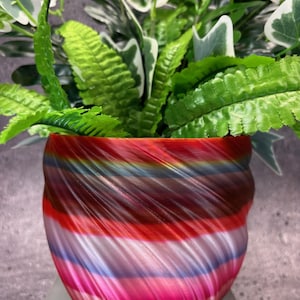 Op de afbeelding: Een kleurrijke, swirl-vormige plantenpot met een kleurenschema van rood, roze, blauw en paars. Een groene, bladerige plant groeit in de pot.