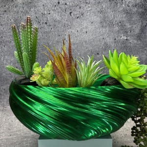 May include: A decorative green planter with a textured, swirling design, filled with artificial succulents and cacti. The arrangement includes a tall cactus, and several other types of succulents. The planter sits on a small, light-coloured base. The overall effect is a striking display.