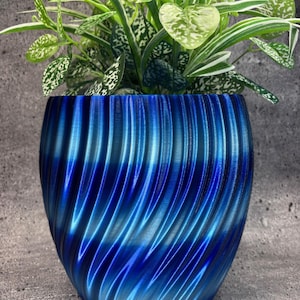 Puede incluir: Una maceta azul con un patrón en espiral contiene una planta verde con hojas moteadas de blanco. La maceta tiene un acabado brillante con tonos azules, creando un contraste visual con el follaje de la planta. El diseño de la maceta añade un toque moderno.