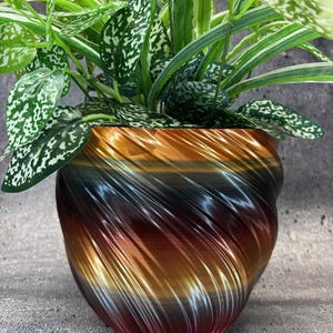 May include: A round plant pot with a metallic, multi-colored gradient finish transitioning from copper and bronze to dark charcoal. The pot features a distinct spiraling, ribbed texture and contains a lush green plant with variegated leaves.