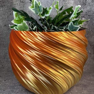 May include: A decorative, swirling planter in shades of gold and orange, holding green and white leafy plants. The planter has a textured, ridged surface, creating a dynamic visual effect. The overall design is modern and eye-catching.