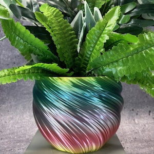 Op de afbeelding: Een kleurrijke bloempot met een wervelend ontwerp, gevuld met een assortiment groene kunstplanten. De pot loopt over van rood naar blauw naar groen, wat een opvallend visueel contrast creëert. De planten hebben diverse bladvormen en texturen.