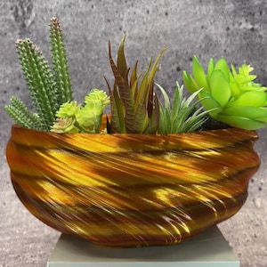 May include: A decorative, gold-coloured planter with a textured, swirling design. The planter is filled with a variety of artificial succulents and cacti in shades of green and brown, creating a vibrant display. The planter is sitting on a white surface.