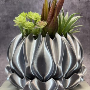 May include: A metallic silver planter with a multi-layered, petal-like exterior design. The planter holds a collection of artificial succulents in shades of light green, dark green, and reddish-brown, creating a dense, textured arrangement.