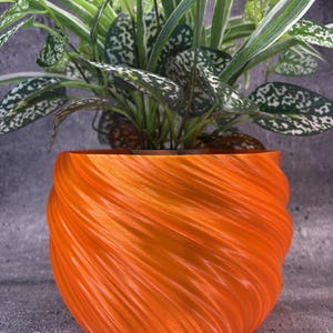 Op de afbeelding: Een feloranje plantenpot met een opvallende, spiraalvormige, geribbelde textuur op het buitenoppervlak. De pot heeft een ronde, bolvormige vorm en bevat een groene plant met wit en groen gevlekt blad dat naar boven uitsteekt.