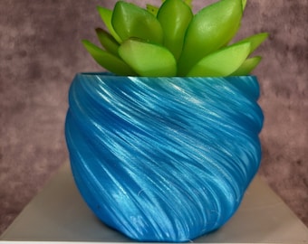 May include: A small, blue, swirled planter pot with a faux succulent plant. The pot is a vibrant, metallic blue with a textured, swirling design. The succulent has green leaves and is centered in the pot. The pot is sitting on a white surface.