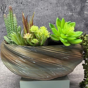 May include: A decorative, oval-shaped planter with a swirling, textured design in shades of brown, gray, and blue. The planter is filled with a variety of artificial succulents and cacti in shades of green and brown. A trailing green plant hangs over the side.