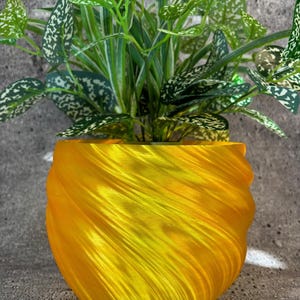 May include: A yellow swirl planter with a green leafy plant inside. The planter is made of a smooth, shiny material and has a unique, swirling design.