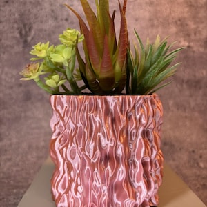 Puede incluir: Una maceta decorativa con un exterior texturizado rosa y naranja, que contiene una variedad de suculentas artificiales. Las suculentas presentan hojas verdes, amarillas y rojizas, creando un contraste vibrante.