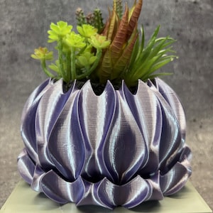 Peut inclure: Un pot décoratif violet et argenté imprimé en 3D avec un design unique en forme de pétale. Le pot est rempli d'une variété de succulentes artificielles dans des tons de vert, de marron et d'orange. Le pot repose sur une surface vert clair.