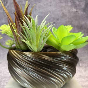 Pode incluir: Um vaso em forma de taça de cor castanho-escuro com um padrão em espiral contendo várias suculentas artificiais. O arranjo inclui folhas altas e pontiagudas de cor castanho-avermelhada e plantas de cor verde.