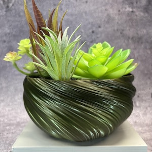 Op de afbeelding: Een donker olijfgroene plantenbak met een wervelende, geribbelde textuur met een arrangement van kunstmatige vetplanten. De planten omvatten een heldere limoengroene rozet, een stekelige lichtgroene plant en langere, slanke roodbruine bladeren.