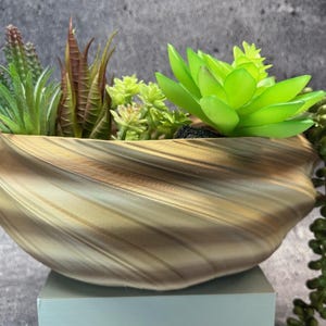 May include: A decorative planter with a swirling, striped pattern in shades of brown and gold. The planter is filled with a variety of artificial succulents in shades of green and brown, creating a natural aesthetic. The planter sits on a light blue surface.