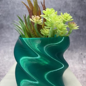 May include: A teal-colored, 3D-printed decorative planter with a wavy design, filled with artificial succulents and greenery. The planter has a unique, twisted shape and is filled with various shades of green and brown faux plants.