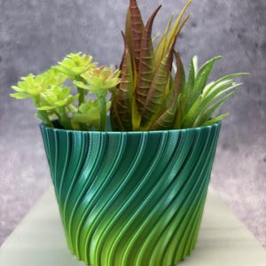 Puede incluir: Una maceta decorativa con degradado verde y amarillo con un diseño en espiral, llena de una variedad de suculentas artificiales. La maceta está sobre una superficie blanca y el fondo es gris texturizado.