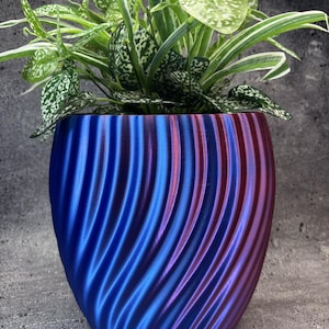 Op de afbeelding: Een plantenpot met een metallic, iriserende afwerking die verschuift tussen diepblauwe en paarse tinten. De buitenkant heeft een verticale, spiraalvormige ribbeltextuur. Een groene en witte bonte plant staat in de ronde houder.