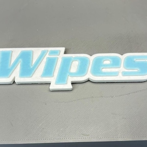 Puede incluir: Un letrero blanco y azul claro con la palabra "Wipes" en azul claro y contorno blanco, colocado sobre una superficie gris. El letrero es un objeto impreso en 3D.
