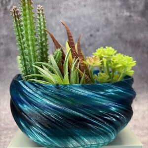 May include: A decorative planter with a swirling blue and teal design, filled with a variety of artificial succulents and cacti. The arrangement includes green cacti, brown and yellow accents, and vibrant green succulents. The planter sits on a white base.