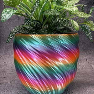 Op de afbeelding: Een kleurrijke, iriserende plantenpot met een draaiende print in tinten roze, groen, blauw en oranje. De pot is gevuld met een groene plant met witte markeringen op de bladeren.