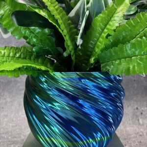 Op de afbeelding: Een arrangement van kunstplanten in een decoratieve, iriserende blauwgroene vaas. De vaas heeft een uniek, wervelend ontwerp. De plant heeft weelderig, groen blad, wat een contrast vormt met de kleuren van de vaas.