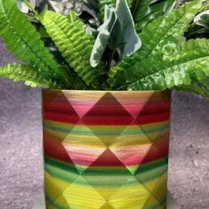 Può includere: Un vaso da fiori geometrico colorato con un motivo a strisce rosso, rosa, verde e giallo. Il vaso è realizzato in un materiale lucido e ha una forma esagonale.