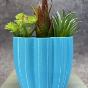 Op de afbeelding: Een kleine, lichtblauwe, geribbelde bloempot gevuld met kunstmatige vetplanten. De vetplanten omvatten groene, bruine en geelgroene planten. De pot is ongeveer 10 cm hoog.