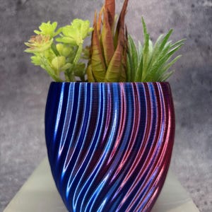 Op de afbeelding: Een kleine, decoratieve plantenbak met een blauw en rood wervelend ontwerp. De plantenbak is gevuld met een verscheidenheid aan kunstmatige vetplanten in groen- en bruintinten. De pot is ongeveer 10 cm hoog.