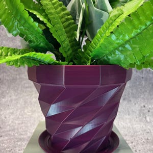 May include: A decorative, geometric-patterned, plum-colored plant pot with a matching saucer. The pot is filled with vibrant green artificial plants. The pot's design features a series of angled, diamond-shaped facets, adding a modern touch to the plant container.