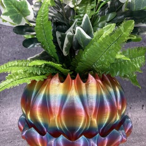 May include: A multi-colored, iridescent planter with a scalloped, petal-like exterior design. The planter features a gradient of rainbow hues including red, orange, yellow, green, and blue. It is filled with a lush arrangement of green foliage.