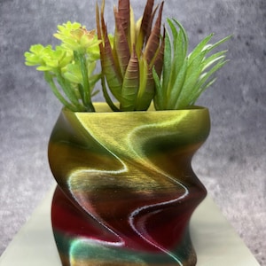 Op de afbeelding: Een decoratieve plantenbak met een metallic afwerking en een uniek, gedraaid ontwerp. De plantenbak is gevuld met een verscheidenheid aan kunstmatige vetplanten in groen, bruin en geel. De kleuren van de plantenbak gaan over van rood naar groen naar goud.